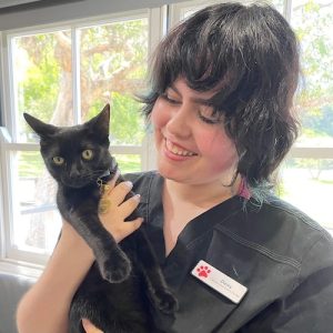 A young woman wearing black scrubs smiles at the black cat she is holding.