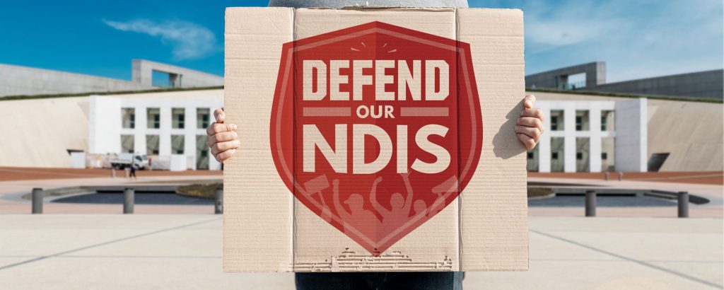 Person standing in front of Parliament House in Canberra holds a placard saying "Defend Our NDIS".
